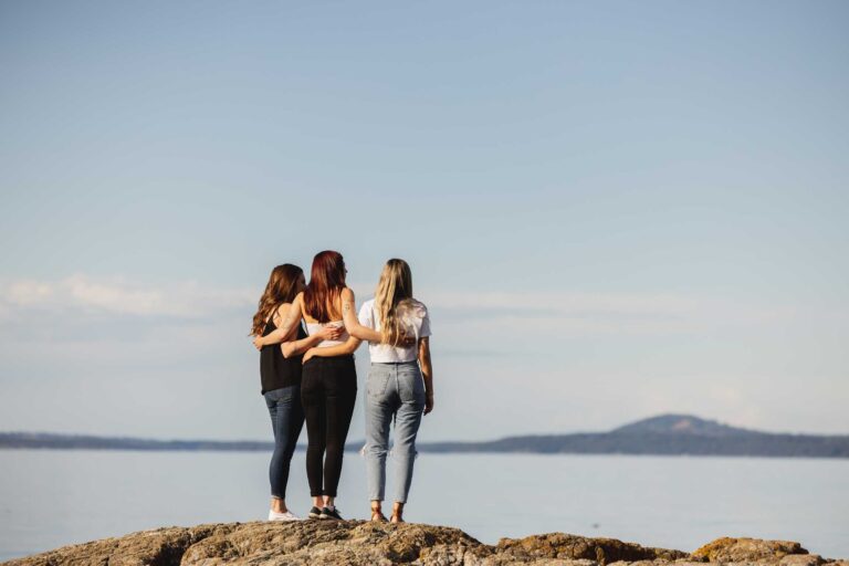 Three women embracing each other standing on a rock looking at the ocean