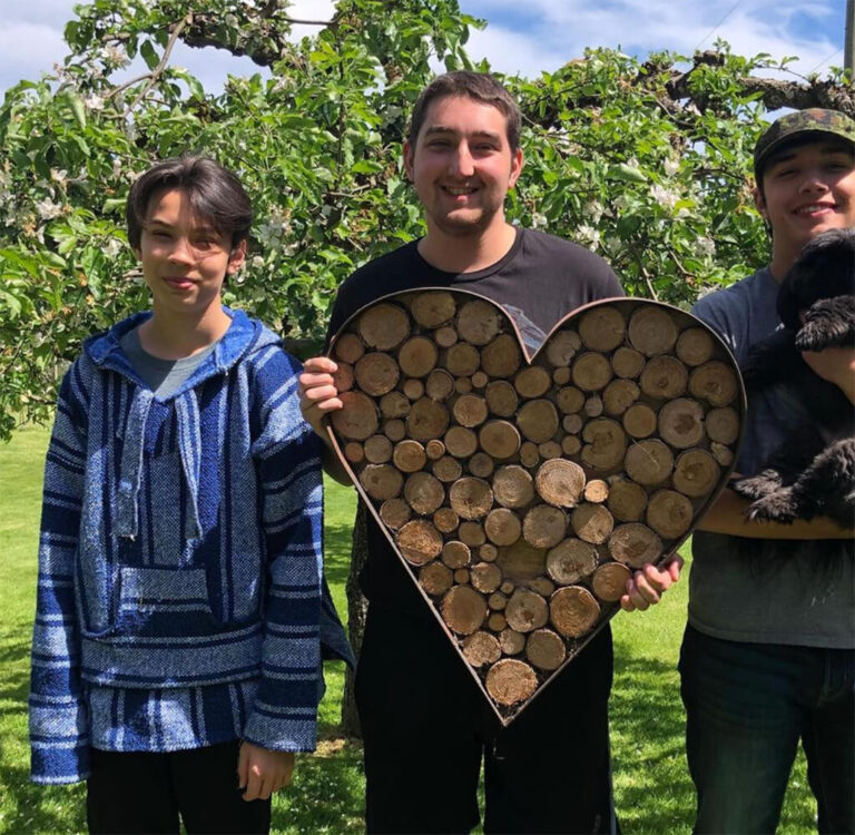 Three brothers smiling and standing in front of a tree, while one is holding a large heart made from metal and small logs of wood as the filler, one of the other boys is holding a puppy