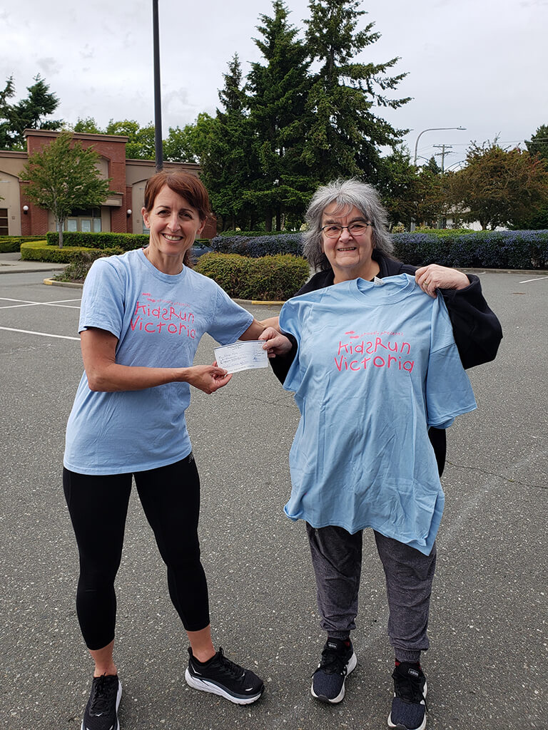 Two women posing for a photo, standing in a parking lot while one is wearing a Kids Run Victoria t-shirt handing another woman a cheque and the same t-shirt