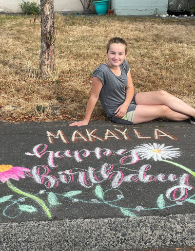 A young girl sitting on grass in front of a pavement path with chalk drawing