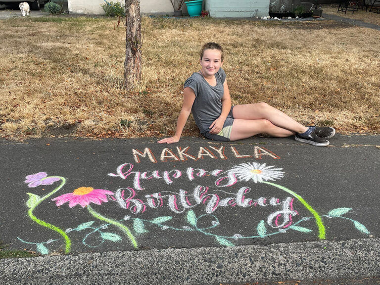 A young girl sitting on grass in front of a pavement path with chalk drawing